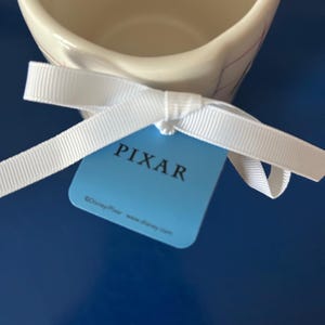 May include: A white ceramic mug with a blue and white design. The mug has a white ribbon tied around it with a blue tag that says "PIXAR" and "&copy;Disney Pixar www.disney.com".