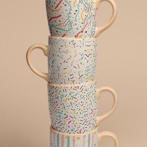 May include: A stack of four ceramic mugs with different colorful geometric patterns. The mugs are stacked on top of each other, with the top mug being a light blue and white striped mug with gold stars.