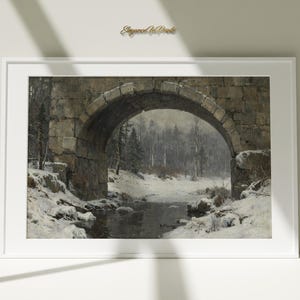 May include: A framed landscape print depicts a stone bridge arching over a snowy stream. The artwork features a winter scene with a forest in the background, showcasing muted tones of gray, brown, and white.