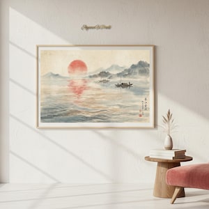Può includere: Un dipinto ad acquerello incorniciato raffigura un sereno paesaggio acquatico con un grande sole rosso che tramonta sull'acqua. Due piccole barche sono sull'acqua e montagne sullo sfondo. Il dipinto ha una cornice in legno chiaro.