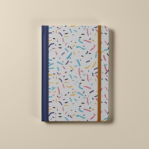 May include: A white notebook with a colorful confetti pattern and a blue spine. The notebook has a brown elastic band closure.