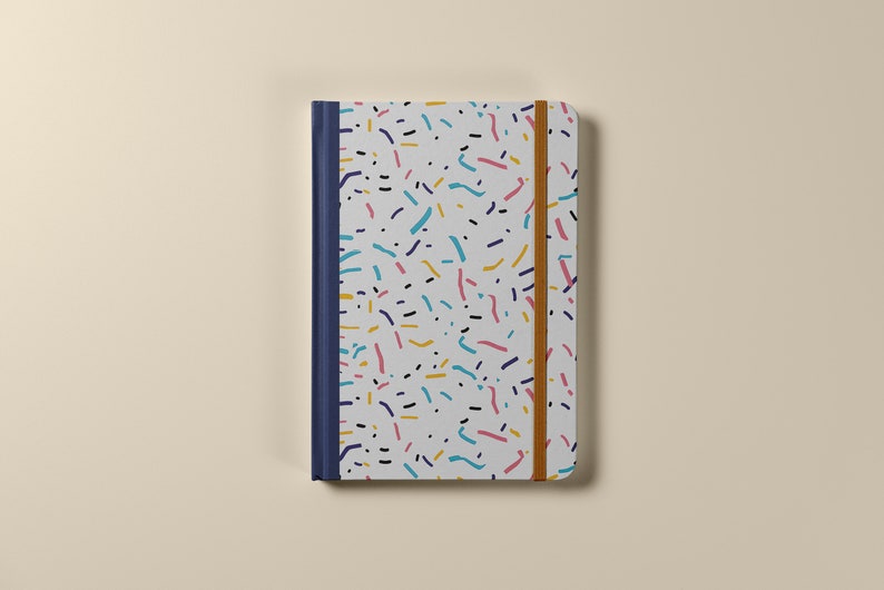 May include: A white notebook with a colorful confetti pattern and a blue spine. The notebook has a brown elastic band closure.