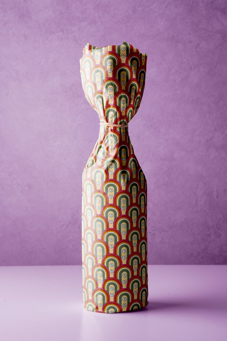 Pode incluir: Uma garrafa embrulhada em papel colorido com um padr&atilde;o de arcos-&iacute;ris e flores brancas. O papel est&aacute; amarrado com um el&aacute;stico.