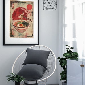 May include: A framed print of a bowl of ramen noodles with a red sun and Japanese text in the background. The print is hanging on a white wall above a white chair with a gray pillow.