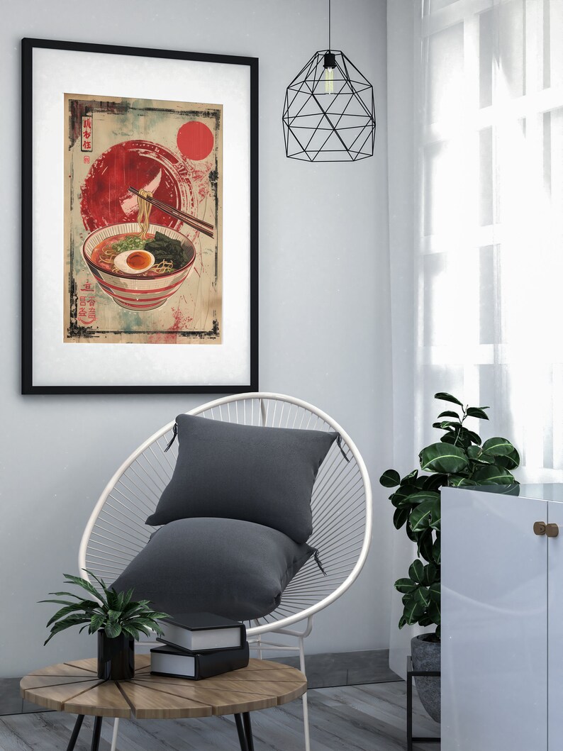 May include: A framed print of a bowl of ramen noodles with a red sun and Japanese text in the background. The print is hanging on a white wall above a white chair with a gray pillow.
