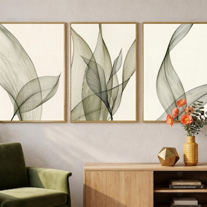 Könnte beinhalten: Drei gerahmte Kunstdrucke mit abstrakten botanischen Designs in Grün- und Grautönen. Das Kunstwerk wird auf einem cremefarbenen Hintergrund in goldenen Rahmen präsentiert. Die Drucke sind über einem Holzschrank angeordnet, mit einem grünen Sessel und dekorativen Gegenständen im Vordergrund.