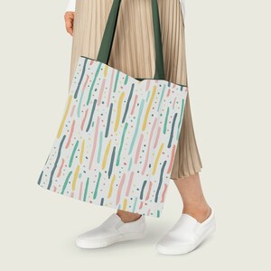 May include: A white tote bag with a colorful abstract pattern of lines and dots. The bag has green straps and is being held by a person wearing a beige skirt.
