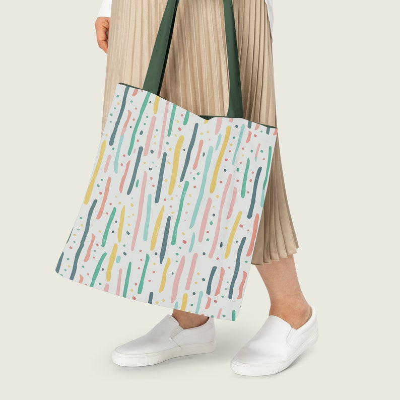 May include: A white tote bag with a colorful abstract pattern of lines and dots. The bag has green straps and is being held by a person wearing a beige skirt.