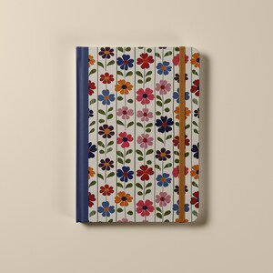 Pu&ograve; includere: Un quaderno bianco con un motivo floreale in tonalit&agrave; di rosa, rosso, blu, arancione e verde. Il quaderno ha una rilegatura blu e una chiusura elastica marrone.