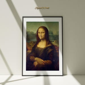 Könnte beinhalten: Gerahmter Kunstdruck der Mona Lisa mit einem humorvollen Element. Das berühmte Porträt zeigt ein kleines, weißes Objekt auf der Nase. Das Kunstwerk ist in einem schwarzen Rahmen vor einer weißen Wand mit Schatten.