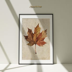 Può includere: Stampa incorniciata con una foglia d'acero dai colori autunnali, dal rosso al marrone dorato. La foglia è posta su uno sfondo beige strutturato, all'interno di una cornice nera. L'immagine è presentata su una superficie bianca.