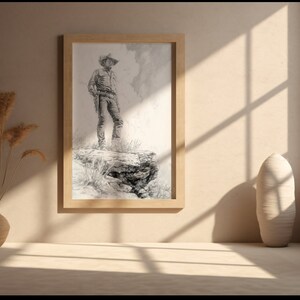 May include: A framed black and white drawing of a cowboy standing on a rock formation, looking out at a distant landscape. The drawing is in a wooden frame and is hanging on a beige wall.