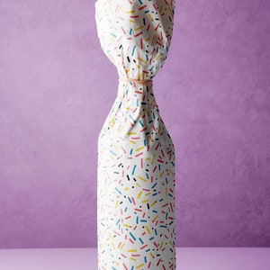 May include: A white bottle wrapped in colorful patterned paper with a rubber band around the neck. The paper has a confetti-like pattern with black, yellow, blue, and red lines and dots.