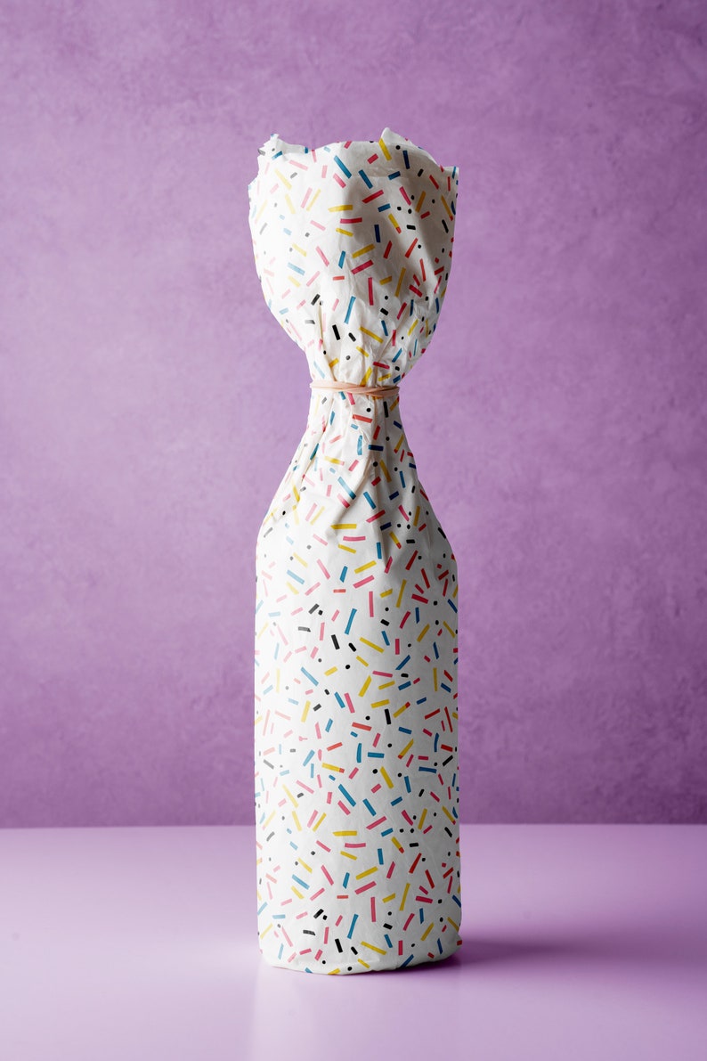 May include: A white bottle wrapped in colorful patterned paper with a rubber band around the neck. The paper has a confetti-like pattern with black, yellow, blue, and red lines and dots.
