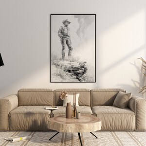 May include: A black and white drawing of a cowboy standing on a rock, framed in a black frame and hanging on a white wall above a beige sofa.