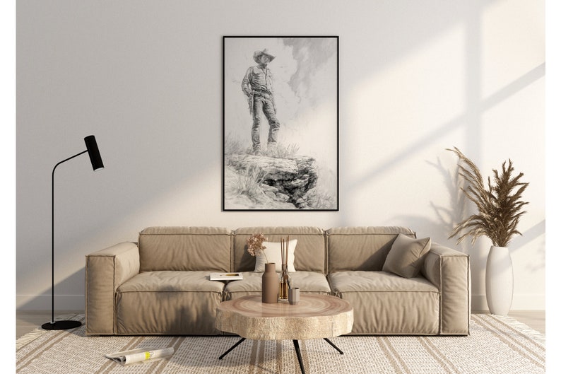 May include: A black and white drawing of a cowboy standing on a rock, framed in a black frame and hanging on a white wall above a beige sofa.