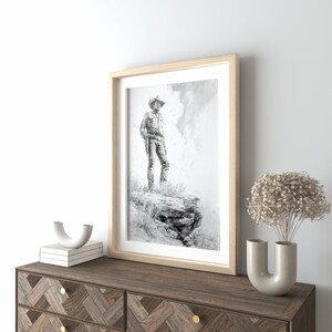 May include: A framed black and white drawing of a cowboy standing on a rocky cliff. The cowboy is wearing a hat and holding a gun. The drawing is set against a cloudy sky.