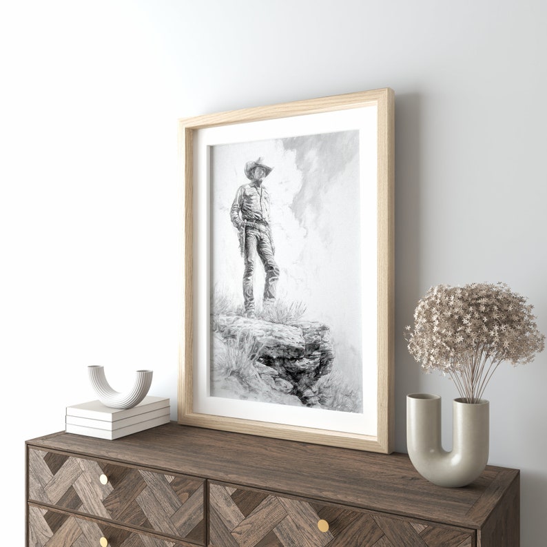 May include: A framed black and white drawing of a cowboy standing on a rocky cliff. The cowboy is wearing a hat and holding a gun. The drawing is set against a cloudy sky.