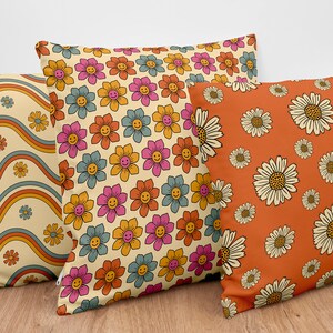 Pode incluir: Tr&ecirc;s almofadas decorativas com padr&otilde;es diferentes de inspira&ccedil;&atilde;o retro. Uma almofada apresenta um padr&atilde;o de listras onduladas em tons de laranja, azul e amarelo. Outra almofada tem um padr&atilde;o repetitivo de flores rosa, azuis e laranja com carinhas sorridentes. A terceira almofada tem um padr&atilde;o repetitivo de margaridas brancas sobre um fundo laranja.