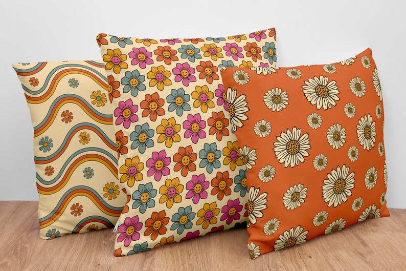 Pode incluir: Tr&ecirc;s almofadas decorativas com padr&otilde;es diferentes de inspira&ccedil;&atilde;o retro. Uma almofada apresenta um padr&atilde;o de listras onduladas em tons de laranja, azul e amarelo. Outra almofada tem um padr&atilde;o repetitivo de flores rosa, azuis e laranja com carinhas sorridentes. A terceira almofada tem um padr&atilde;o repetitivo de margaridas brancas sobre um fundo laranja.