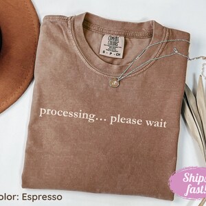 May include: Espresso-colored t-shirt with the text "processing... please wait" in white. The shirt has a crew neck and a small tag near the collar. A silver necklace with a pendant is draped across the shirt. A brown hat is visible in the background.