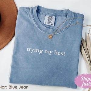 Puede incluir: Una camiseta azul vaquero con la frase "trying my best" impresa en blanco. Un collar dorado con un colgante está drapeado sobre la camiseta. La camiseta está doblada y se muestra con un sombrero marrón y hojas secas.