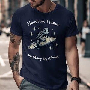Può includere: Una t-shirt blu navy con una grafica bianca di una tartaruga che cavalca un razzo con stelle e il testo "Houston, I Have So Many Problems".