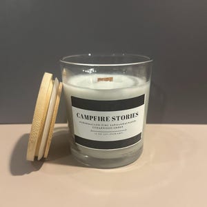 May include: A glass candle jar with a wooden lid. The candle is white and has a black label that reads "Campfire Stories" with the scent notes "Marshmallow, Pine, Sapsi, Sandalwood, Cedarwood, Amber" and "10 oz (283 grams)".