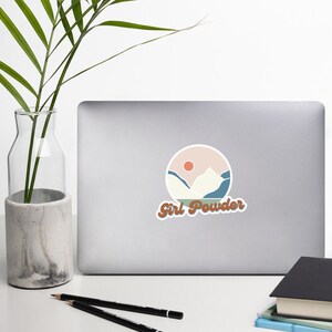 Può includere: Un adesivo con un design di paesaggio montano in stile retrò. Le montagne sono bianche con accenti blu, e il cielo è rosa con un sole rosso. Il testo "Girl Powder" è in marrone con un font retrò.