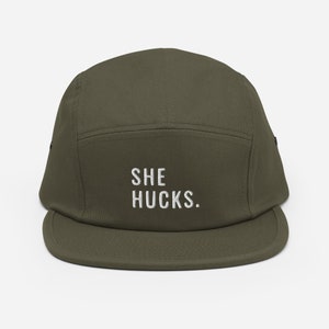Può includere: Cappellino a cinque pannelli verde oliva con visiera piatta. Il cappellino presenta la scritta ricamata bianca "SHE HUCKS" centrata sul pannello frontale. Il cappello è realizzato in tessuto resistente e ha un design strutturato.