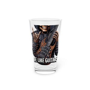 May include: A clear pint glass featuring a detailed illustration of a skeleton in a leather jacket holding a guitar. The skeleton is making a hand gesture. The text "DIE LIKE GUITARS" is printed on the glass.
