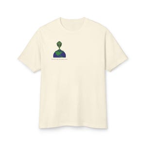 Puede incluir: Camiseta color crema con una cabeza de alienígena verde sobre un gráfico de la Tierra azul y verde. El texto "earthing_invaders.com" está impreso debajo. La camiseta tiene mangas cortas y cuello redondo.