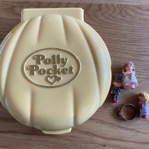 Può includere: Un playset Polly Pocket vintage a forma di conchiglia, con la scritta "Polly Pocket" in lettere dorate. Include figurine in miniatura di una sposa, uno sposo e un bambino, insieme a un piccolo anello. Il set è su una superficie di legno.