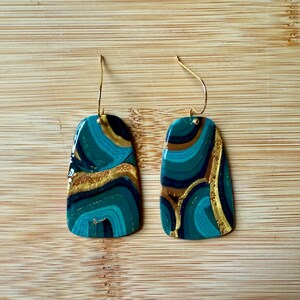 May include: A pair of artisan earrings with a marbled design. The earrings feature a teal, gold, and black pattern on a rectangular shape with gold-colored ear wires. The earrings are displayed on a wooden surface.
