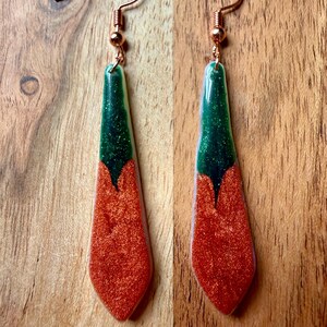 May include: A pair of handmade earrings with copper-colored ear wires. The earrings have a long, teardrop shape, with a glittery green top and a glittery orange-red bottom. The earrings are displayed on a wooden surface.