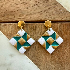 May include: A pair of dangle earrings featuring a diamond-shaped pendant with a geometric pinwheel pattern. The design consists of white, teal, and metallic gold segments. Each earring is attached to a small, round, polished gold-tone stud.