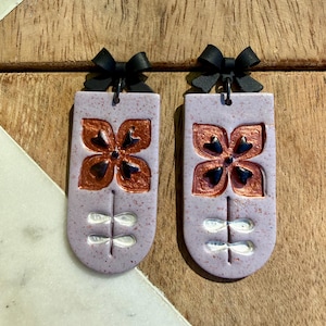 May include: A pair of lavender-colored earrings with a floral design. Each earring features a copper-colored flower with black accents and a white stem with two pairs of leaves. A black bow tops each earring.