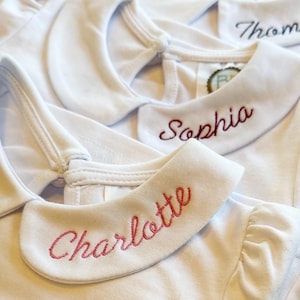 May include: Close-up of several white baby shirts with Peter Pan collars. Each collar is embroidered with a different name in red thread: Charlotte, Sophia, and Thomas. The shirts are made of a soft, light-colored fabric.