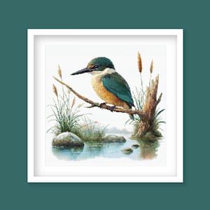 Op de afbeelding: Een ingelijste kruissteek van een ijsvogel die op een tak boven een waterpartij zit. De vogel heeft een groene kop, turkooizen vleugels en een gele borst. Riet en rotsen zijn zichtbaar in het water.