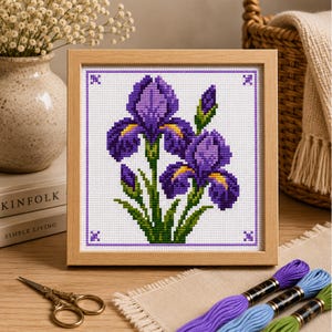 May include: A square cross-stitch embroidery featuring two purple iris flowers with yellow accents and green stems, framed in a light-colored wooden frame. The design is stitched on white fabric with a purple border and small corner motifs.
