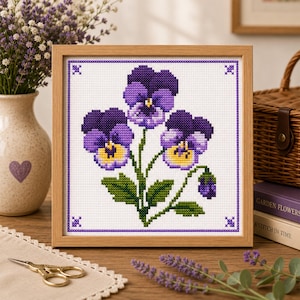 Puede incluir: Un bordado en punto de cruz cuadrado que presenta tres flores de pensamiento moradas y amarillas con hojas verdes, enmarcado en un marco de madera clara. El diseño incluye un borde de punto de cruz morado y cuatro motivos decorativos en las esquinas.