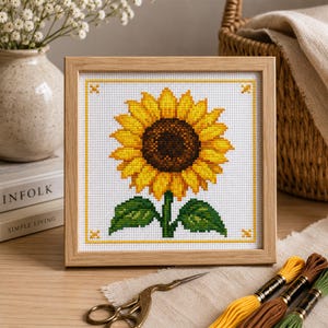 May include: A square wooden frame containing a cross-stitch embroidery of a single sunflower with bright yellow petals, a dark brown center, and two green leaves on a white fabric background, featuring small yellow cross-stitch accents in the corners.