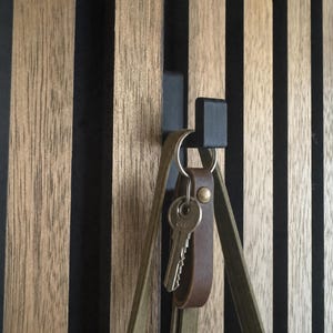 Könnte beinhalten: Ein mattschwarzer Wandhaken aus Metall, montiert auf einer Platte aus vertikalen, hellbraunen Holzlatten mit schwarzen Zwischenräumen. Ein brauner Lederschlüsselanhänger mit einem silbernen Metallschlüssel und ein grüner Stofftaschenhenkel hängen daran.