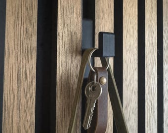 Könnte beinhalten: Ein mattschwarzer Wandhaken aus Metall, montiert auf einer Platte aus vertikalen, hellbraunen Holzlatten mit schwarzen Zwischenräumen. Ein brauner Lederschlüsselanhänger mit einem silbernen Metallschlüssel und ein grüner Stofftaschenhenkel hängen daran.