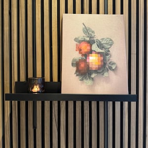 May include: A black shelf displays a framed artwork of pixelated oranges with green leaves. A lit candle in a dark glass holder sits to the left. The background is a wooden wall with vertical stripes.