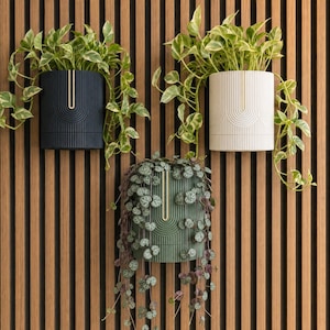May include: Three cylindrical wall-mounted planters in dark blue, off-white, and sage green. Each planter features a vertical ribbed texture with a central U-shaped cutout. Trailing green foliage grows from the top of each container.