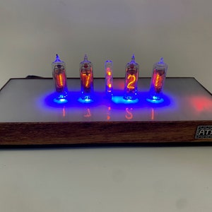 May include: A wooden clock with six red glowing Nixie tubes displaying the time 17:12. The clock has a white face with blue and red LED lights around the tubes. The clock is labeled "ATHNK" on the bottom right.