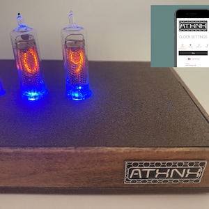 May include: A wooden clock with four red Nixie tubes displaying the number 109. The clock has a black surface and a brown wooden frame with the word "ATHNK" printed on it. The clock is connected to a smartphone app for setting the time.