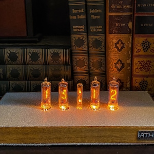 May include: A wooden clock with a white face and five glowing orange Nixie tubes displaying the time 1:51:12. The clock is sitting on a shelf with a row of books behind it. The clock has a small button on the side and the word "ATHNH" printed on the bottom.