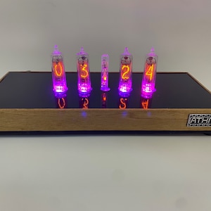 May include: A wooden clock with six clear glass tubes displaying the time as 03:24. The tubes are lit with a purple glow and the numbers are orange. The clock has a black glass top and a wooden base with a silver label that reads "ATHN".